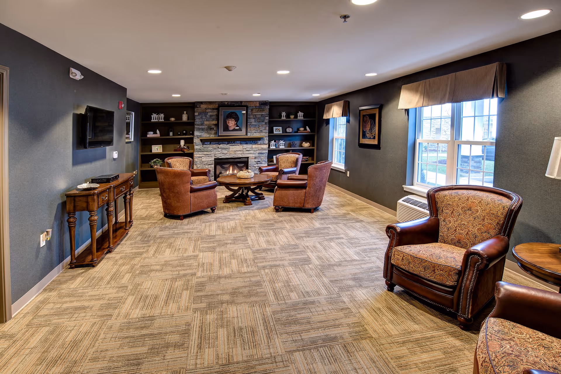 A cozy living room area with four brown leather armchairs arranged around a round wooden coffee table in front of a stone fireplace. The fireplace is flanked by built-in shelves with decorative items. There are two large windows with beige valances on the right wall, allowing natural light to enter. A flat-screen TV is mounted on the left wall above a wooden console table. The room has carpeted flooring and recessed ceiling lights.