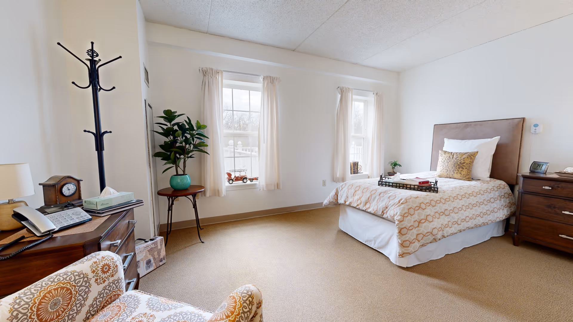 Bright, neatly furnished senior living bedroom with a single bed, nightstands, a chair, windows, and a potted plant.