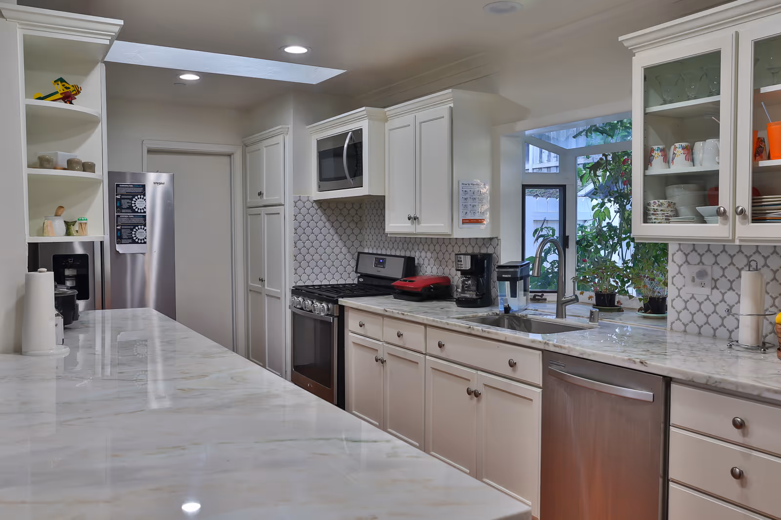 Modern kitchen with white cabinetry, marble countertops, stainless steel appliances including a refrigerator, stove, microwave, and dishwasher. There is a coffee maker and a water filter on the counter, a window above the sink with plants visible outside, and recessed lighting in the ceiling.