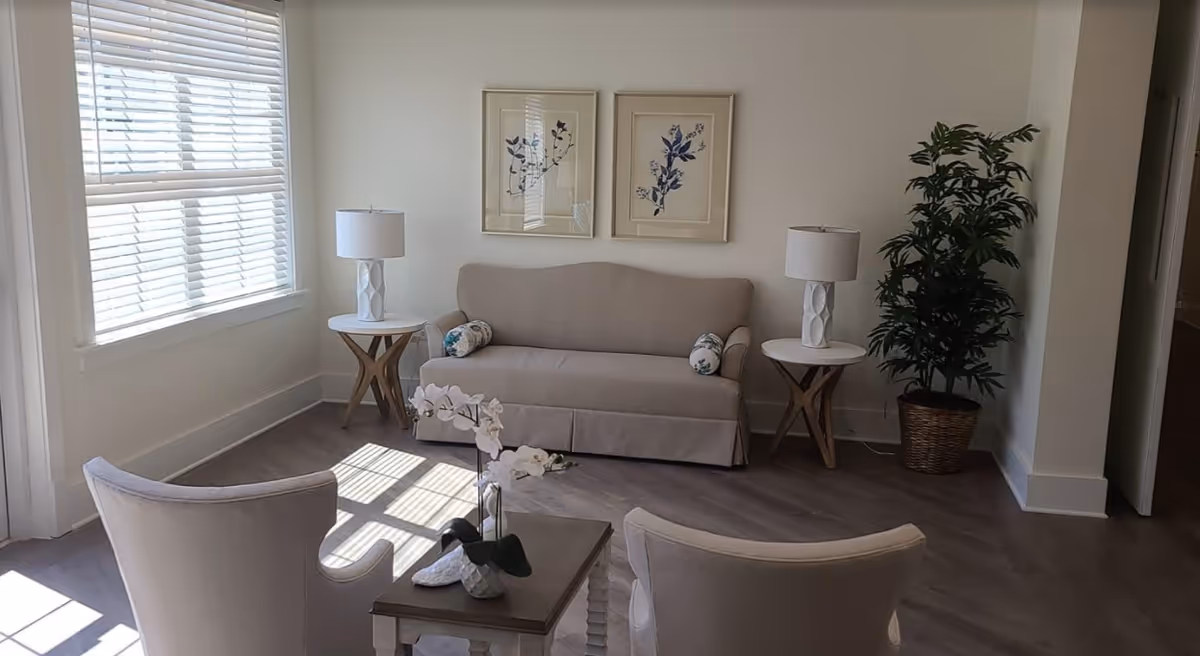 A bright living room with a beige sofa flanked by two white side tables each holding a white lamp. Two framed botanical prints hang above the sofa. In front of the sofa are two beige armchairs and a small wooden coffee table with a white orchid plant. A large window with white blinds lets in natural light, and a tall potted plant is placed in the corner.