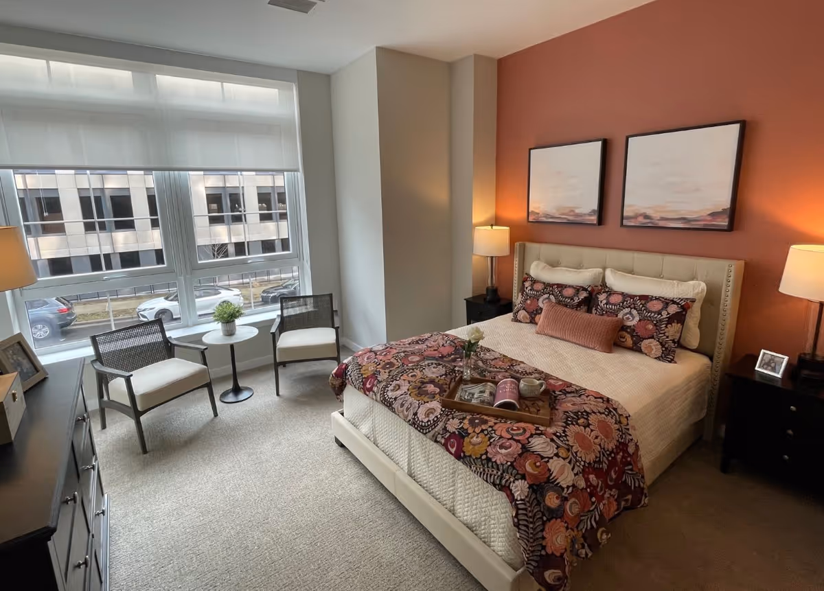 Bright, modern bedroom with a bed against a coral accent wall, bedside tables and lamps, decorative artwork, and two chairs by a large window.