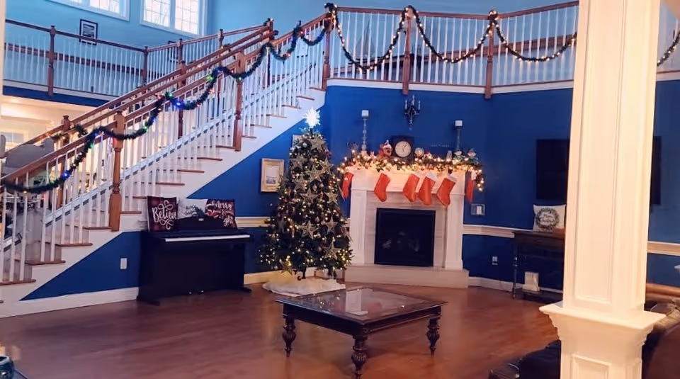 A cozy living room decorated for Christmas with a Christmas tree adorned with lights and ornaments next to a white fireplace with stockings hanging from the mantel. The mantel is decorated with garlands and candles. A staircase with wooden handrails and garlands wrapped around the banister is visible in the background. There is a black piano with festive pillows on the left side and a wooden coffee table in the center of the room. The walls are painted blue with white trim.