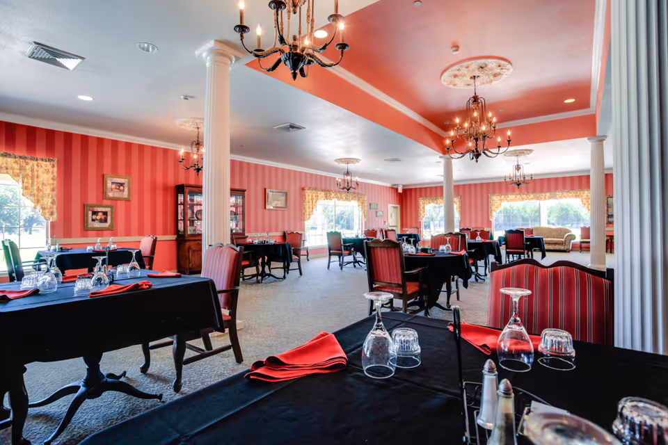 Spacious dining room with red-striped walls, chandeliers, columns, and tables set with black cloths, red napkins, and glassware.