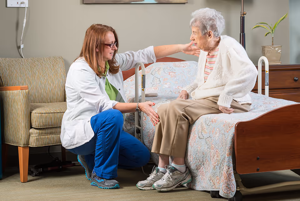A healthcare worker in a white coat and blue pants is kneeling beside an elderly woman sitting on a bed in a room. The worker is gently touching the woman's face and knee, providing support and care. The room has a patterned bedspread, a wooden headboard, a chair, and a small table with a plant.