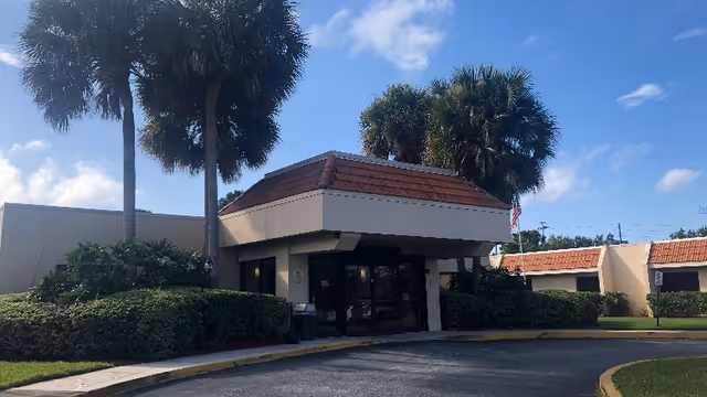 Exterior view of a single-story building with a tiled roof entrance, surrounded by palm trees and bushes under a partly cloudy blue sky.