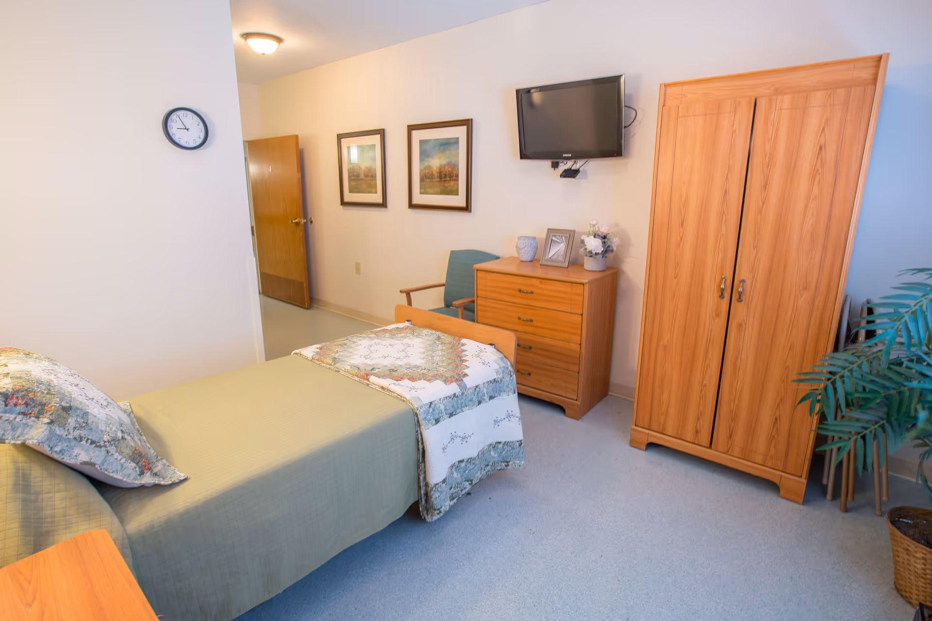 A neatly arranged bedroom in a senior living facility with a single bed covered in a green bedspread and a patterned quilt. The room features wooden furniture including a dresser with decorative items, a wardrobe, and a chair. A flat-screen TV is mounted on the wall above the dresser. Two framed pictures hang on the wall, and a clock is visible near the doorway. There is also a potted plant in the corner.