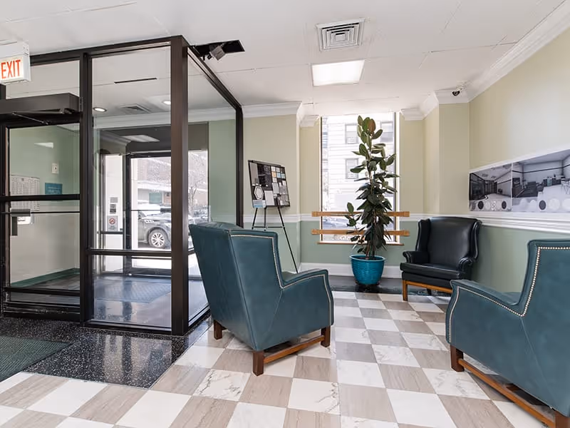 A small seating area inside Commonwealth Apartments featuring three dark green leather armchairs arranged around a potted plant in a blue pot. The floor has a checkered pattern of beige and white tiles. There is a glass entrance door with a black frame on the left side, and a bulletin board on a stand near the window. The walls are painted light green with white trim, and a large window lets in natural light.