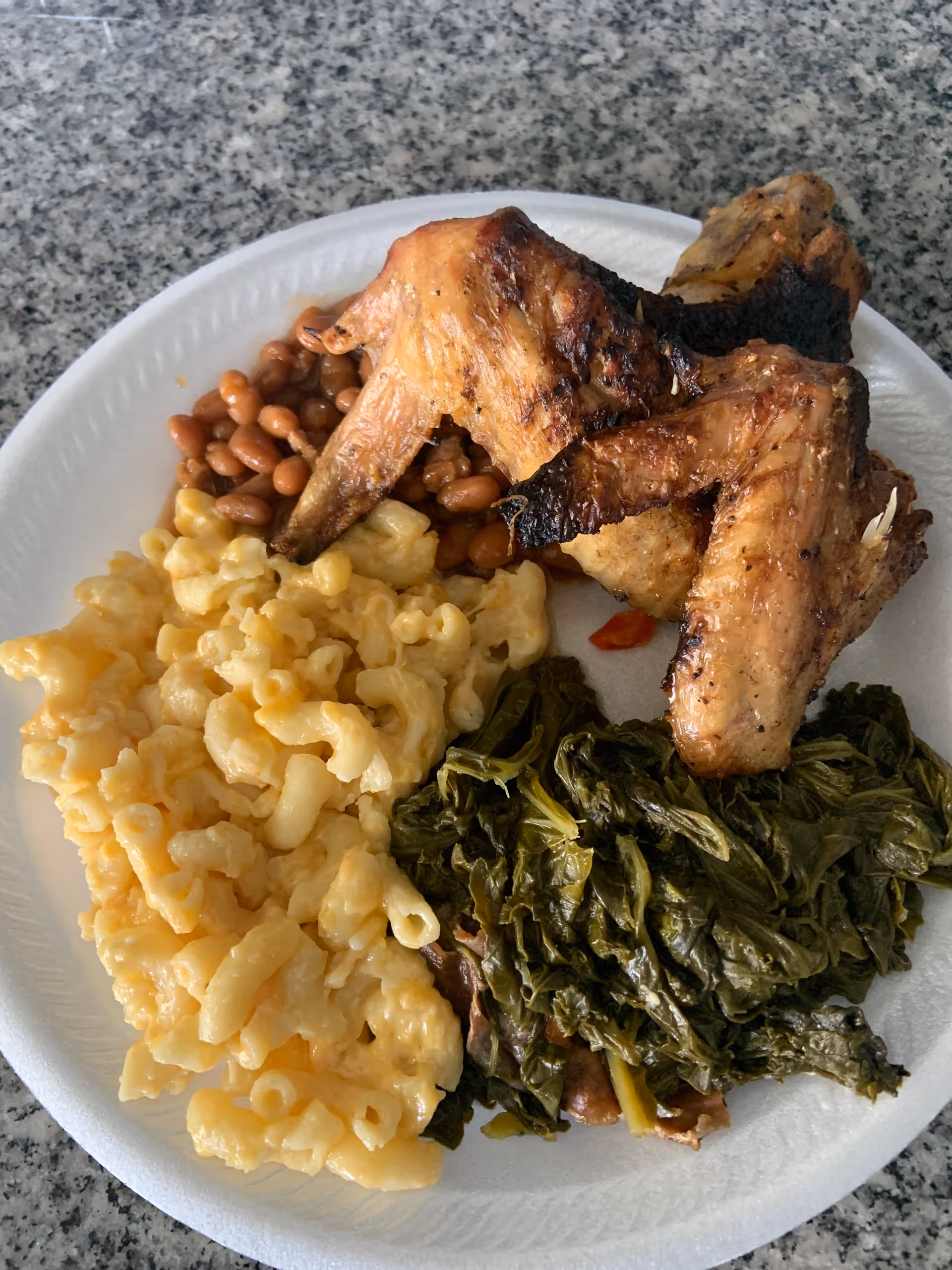 A white disposable plate with a serving of baked beans, macaroni and cheese, cooked greens, and two grilled chicken wings on a granite countertop.