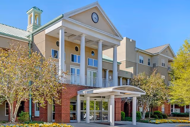 Exterior view of Brookdale University Park building featuring a grand entrance with white columns, a clock on the triangular pediment, and a covered drop-off area. The building has a combination of brick and beige siding with green metal roofing. Trees and landscaping with flowers surround the entrance under a clear blue sky.