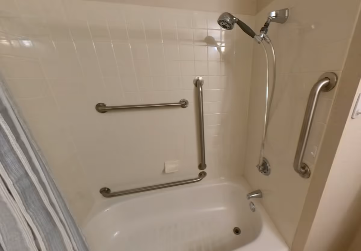 Bathtub with white tiled walls, equipped with three stainless steel grab bars and a handheld showerhead. A striped shower curtain is partially visible on the left side.