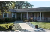 Single-story building with a covered porch, white picket fence, and a concrete walkway leading to the entrance, surrounded by grass and trees.