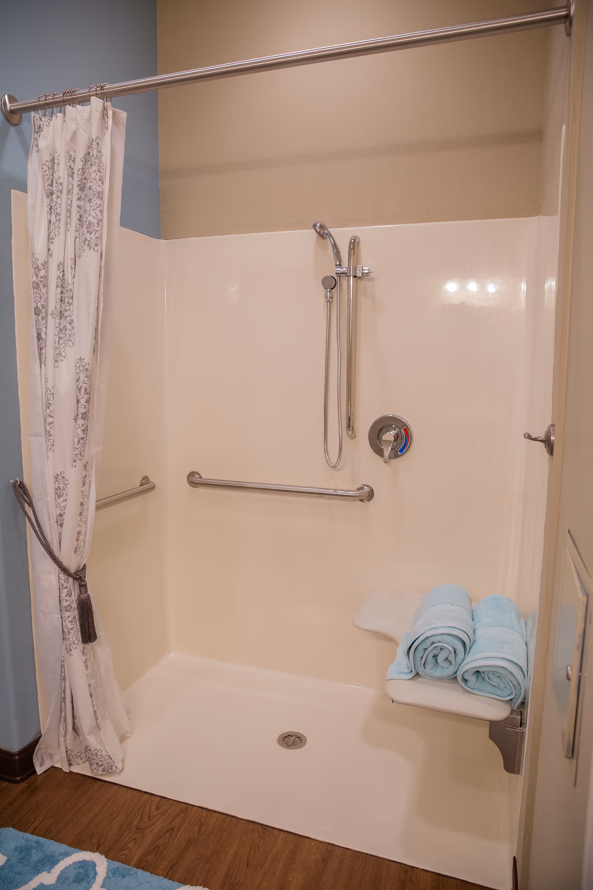 Accessible shower area with a handheld showerhead, grab bars, a fold-down seat with three rolled blue towels on it, and a decorative shower curtain tied back to one side. The shower has a beige wall surround and a wooden floor outside.