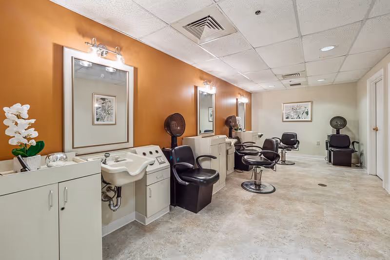 Interior view of a hair salon area with three styling stations, each equipped with a mirror, a black salon chair, and a hair dryer chair. The walls are painted orange and beige, and there is a white cabinet with a white orchid plant on top. The floor is light-colored and the ceiling has recessed lighting.