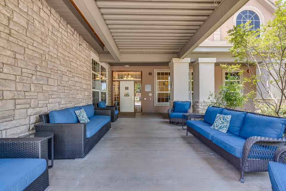 Covered outdoor seating area at Riverstone Terrace Senior Living with wicker furniture featuring blue cushions and patterned pillows, stone wall on the left, and entrance door with windows in the background.