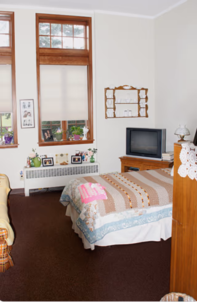 A tidy bedroom with a quilted bed, TV on a wooden dresser, large windows with shades, and personal photos and decorations.