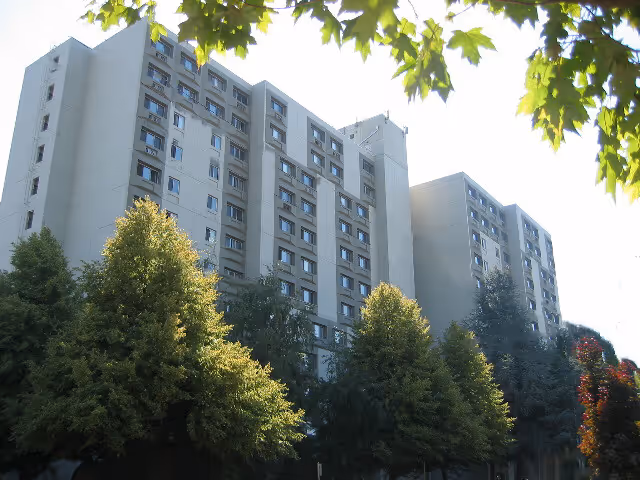 Exterior view of a multi-story residential building with numerous windows, surrounded by tall green trees and leafy branches in the foreground.