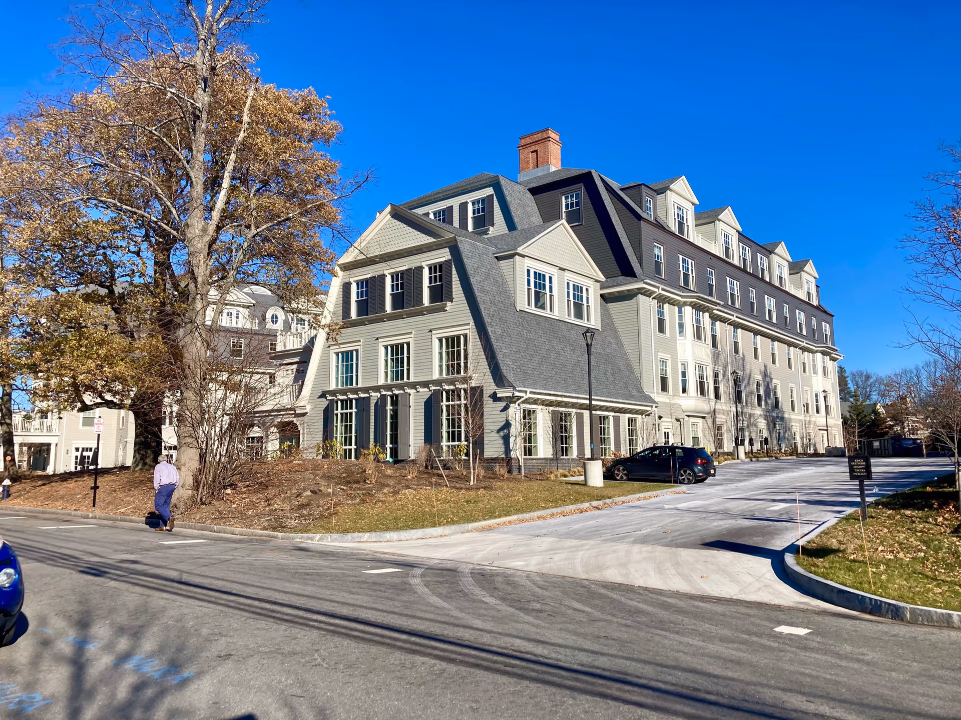 Large multi-story colonial-style building front with a driveway, parked car, and trees under a clear blue sky.