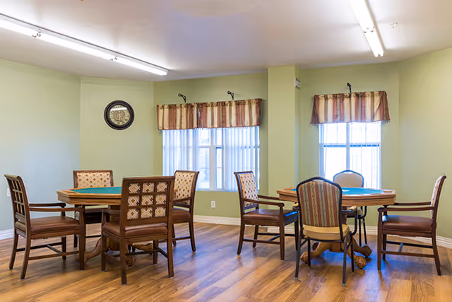 A well-lit room with two round wooden tables, each surrounded by four cushioned chairs with patterned upholstery. The walls are painted light green, and there are two windows with striped valances letting in natural light. A round clock is mounted on the wall above one table. The floor is wood laminate.