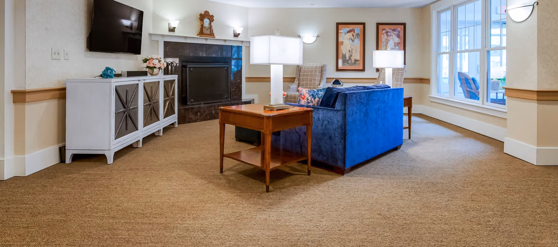 Cozy common room with a blue sofa, wooden side tables and lamps facing a wall-mounted TV and fireplace near large windows.