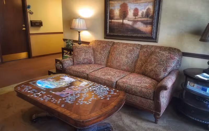 A cozy living room area featuring a patterned three-seat sofa with a matching pillow, a wooden coffee table with a partially completed jigsaw puzzle on it, a side table with a lamp and some magazines, and a framed painting of trees on the wall behind the sofa.