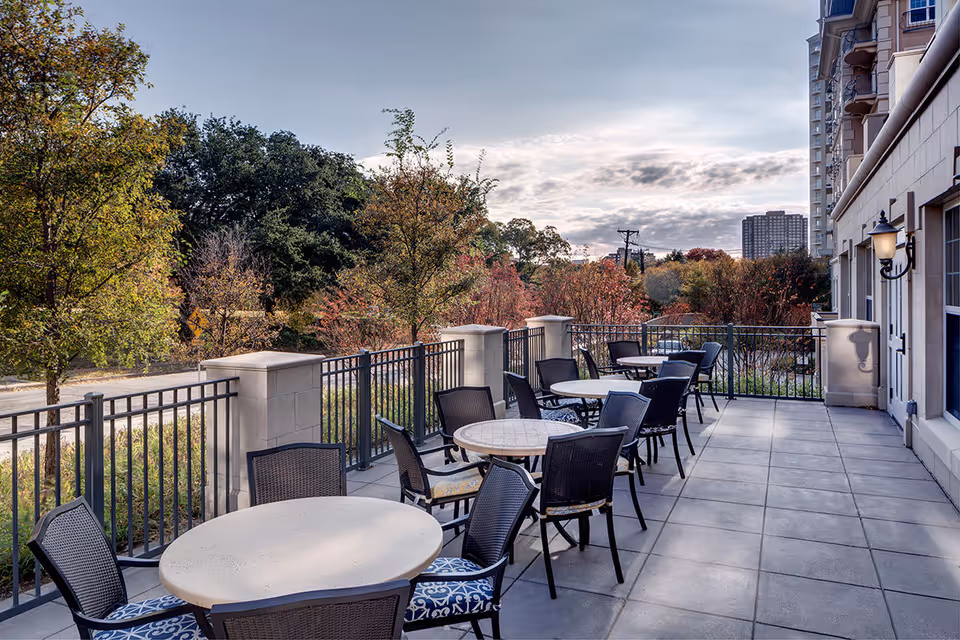 Outdoor terrace with multiple round tables and chairs along a building, fenced overlooking trees and distant city buildings.