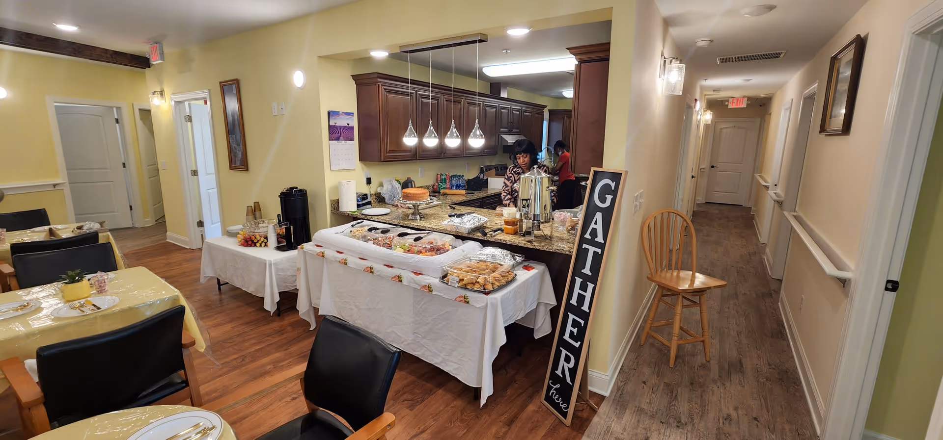 Interior view of a senior care home dining and kitchen area with tables covered in yellow tablecloths and set with plates and utensils. A buffet table with food trays is set up near the kitchen counter where two women are preparing or serving food. A tall wooden sign with the word 'GATHER' leans against the wall near a wooden chair. The hallway adjacent to the kitchen leads to other rooms, with light-colored walls and wood flooring throughout.