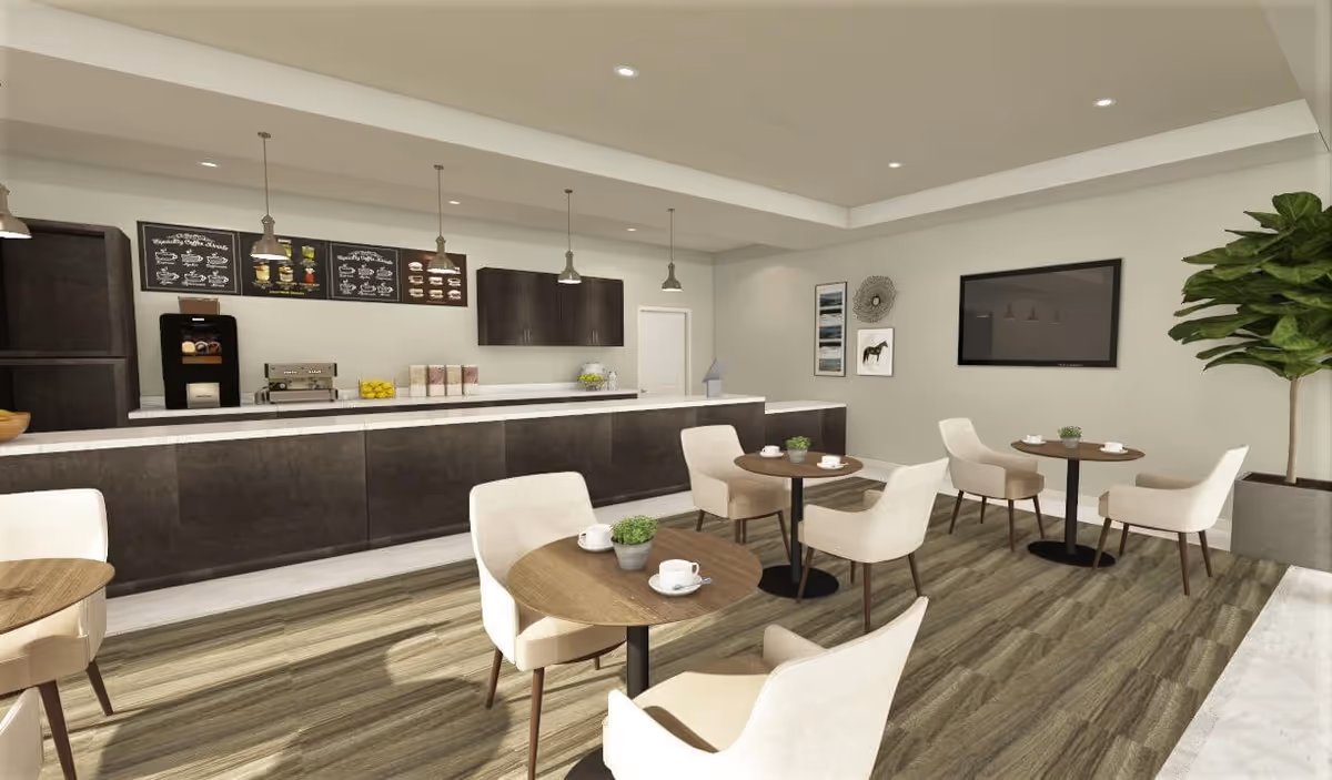 A modern dining area with several round wooden tables and cream-colored upholstered chairs. The room features a long counter with a coffee machine, espresso machine, and various items on display. Above the counter are three pendant lights and menu boards. The walls are decorated with framed pictures and a large flat-screen TV. There is a large potted plant in the corner and wood-patterned flooring.