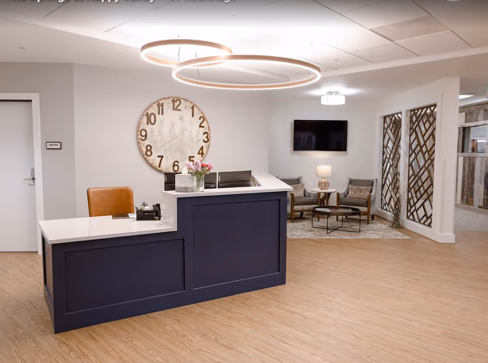 Reception desk and seating area in a senior living facility lobby with a large wall clock and modern circular ceiling lights.