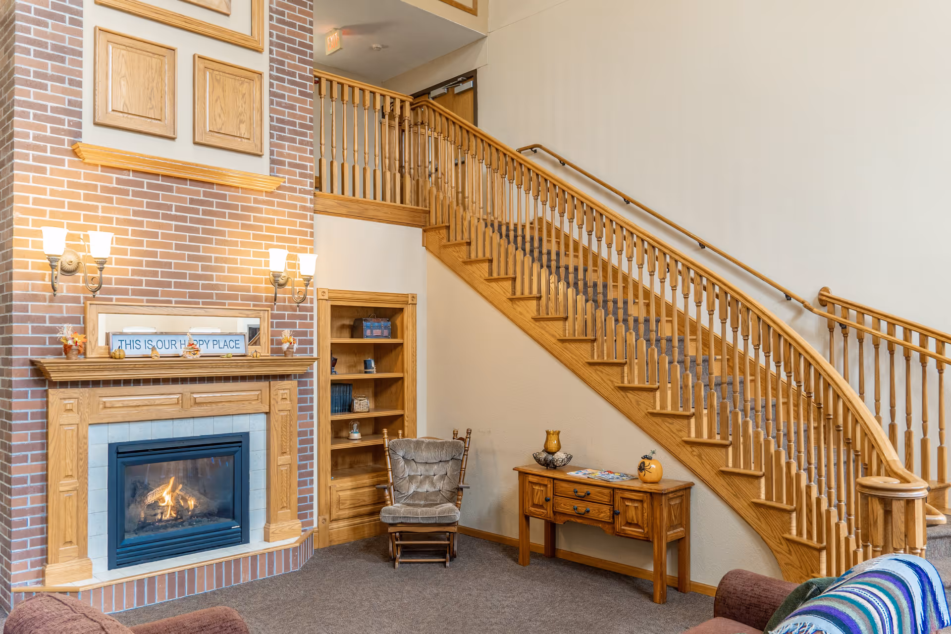 A cozy interior living area featuring a brick fireplace with a wooden mantel, a cushioned rocking chair, a wooden side table with decorative items, and a carpeted staircase with wooden railings leading to an upper floor.
