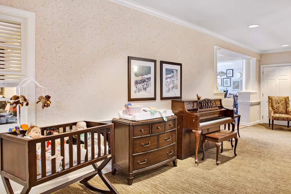 A bright interior common room featuring a wooden baby crib with dolls, a dresser, and an upright piano with seating.