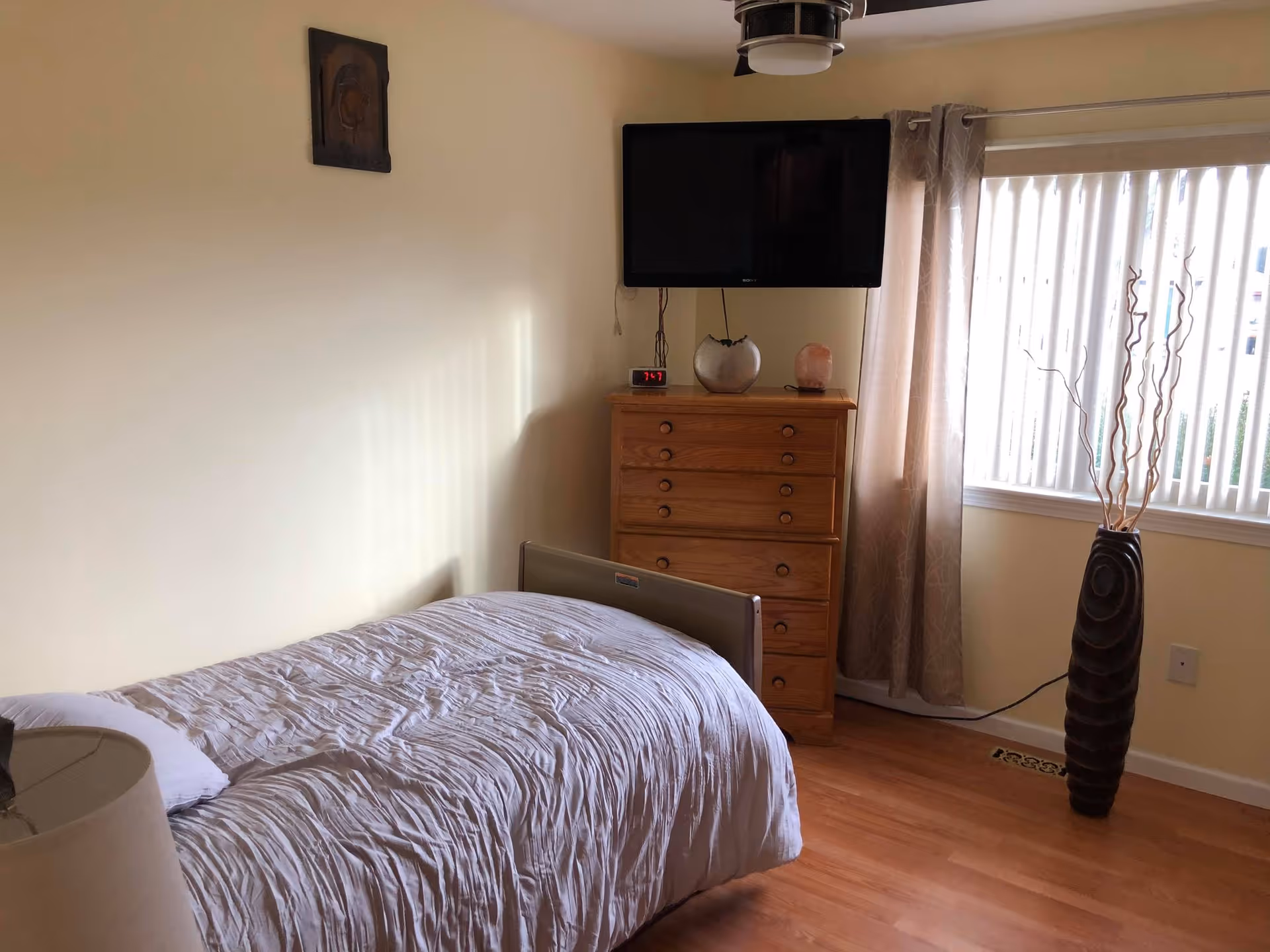 A small bedroom with a single bed covered in a light gray comforter, a wooden dresser with a digital clock, a decorative vase, and a salt lamp on top. A flat-screen TV is mounted on the wall above the dresser. There is a window with vertical blinds and beige curtains, and a tall decorative vase with dried branches stands next to the window. The floor is wooden, and the walls are painted light beige.