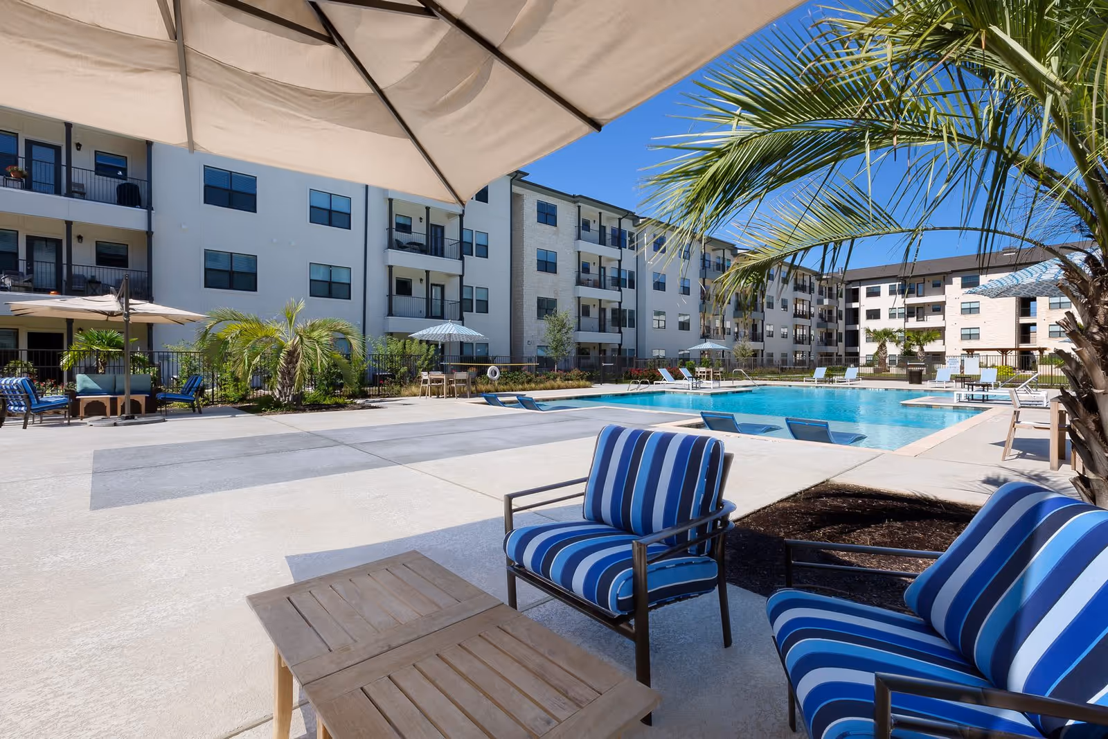 Outdoor pool area at a senior living facility with blue and white striped cushioned chairs, wooden tables, large umbrellas, palm trees, and a multi-story building in the background under a clear blue sky.