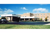 Front exterior of a low-rise brick senior living facility with a covered porte-cochère entrance, a parked car, and a grassy foreground under a blue sky.