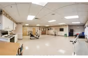 A spacious rehabilitation room with exercise equipment including stationary bikes and parallel bars. The room has a tiled ceiling with fluorescent lighting, beige walls, and a linoleum floor. There are cabinets and a countertop along one wall, and a few chairs are visible. The room appears clean and well-lit, designed for physical therapy activities.