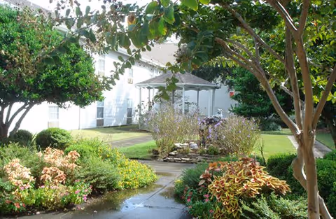A landscaped garden area with a paved walkway leading to a white gazebo surrounded by various green shrubs, flowering plants, and trees. A white building is visible in the background.