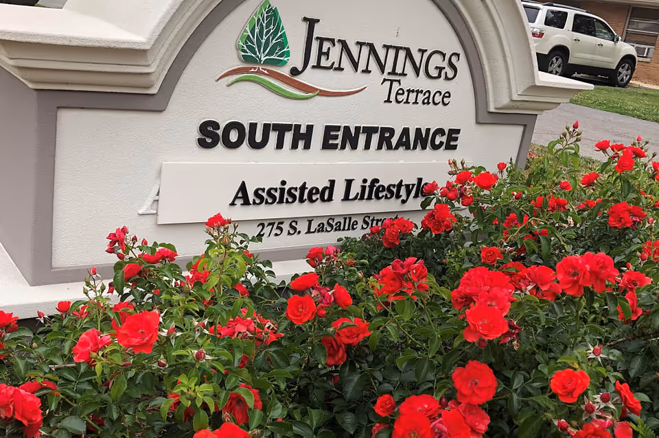 Outdoor sign for Jennings Terrace South Entrance with red flowers in the foreground and a white vehicle parked in the background.