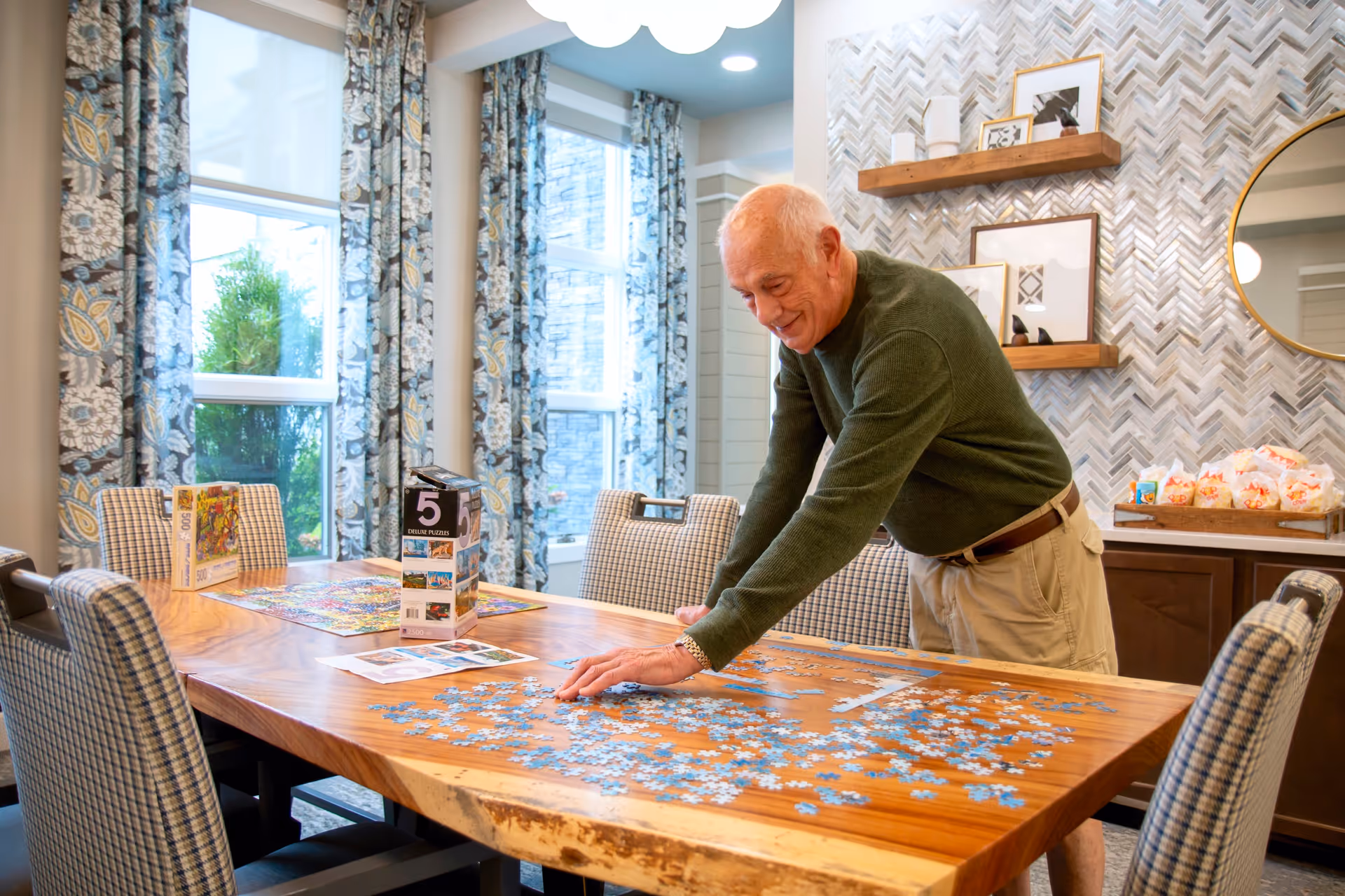 An elderly man wearing a green sweater and beige shorts is standing and working on a jigsaw puzzle on a large wooden table in a well-lit room. The room has patterned curtains on the windows, a decorative wall with shelves holding framed pictures and vases, and a round mirror. There are several chairs with checkered upholstery around the table.