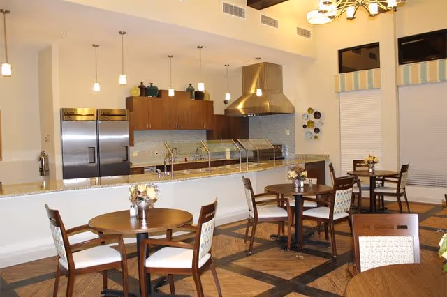 A modern dining area with round wooden tables and chairs arranged neatly. In the background, there is a kitchen area with stainless steel appliances, including refrigerators and a large range hood. Pendant lights hang from the ceiling, and decorative vases are placed on top of the kitchen cabinets. The room has a warm and inviting atmosphere with a combination of wood and neutral tones.