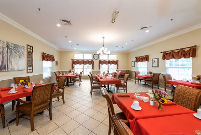 A bright dining room with multiple tables covered in red tablecloths, each set with white cups, glasses, and small flower arrangements. The room has large windows with brown valances, beige walls, tiled floors, and a chandelier hanging from the ceiling. There are framed pictures and a clock on the walls.