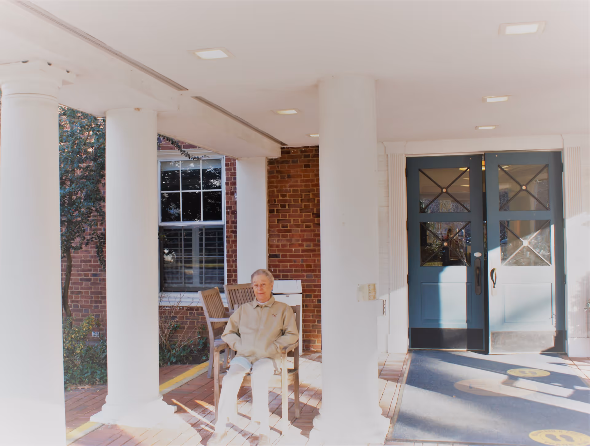 An elderly person sitting on a wooden chair under a covered entrance with white columns at a brick building. There are blue double doors in the background and some wooden chairs stacked behind the person.
