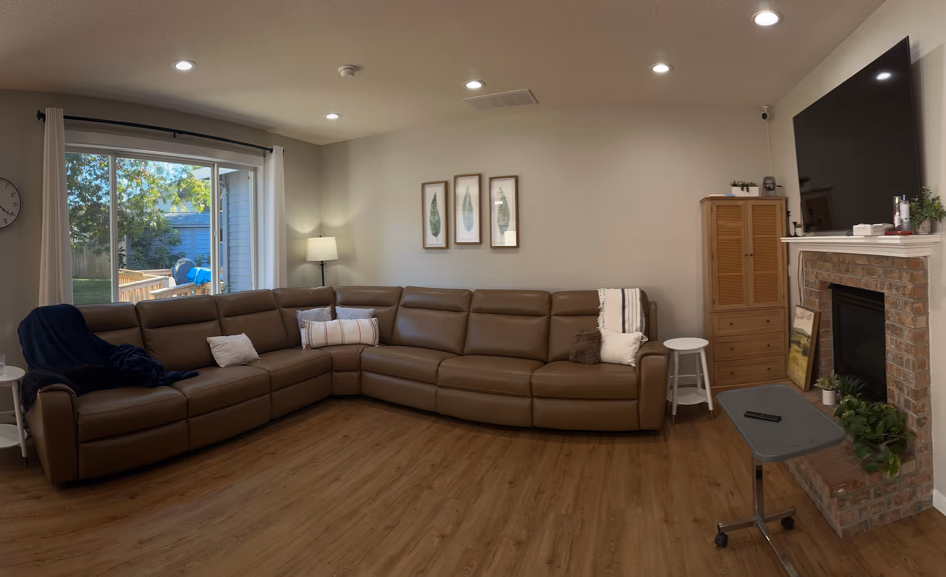Living room with a large brown leather sectional sofa, wood floors, a brick fireplace with wall-mounted TV, and sliding glass doors to the backyard.