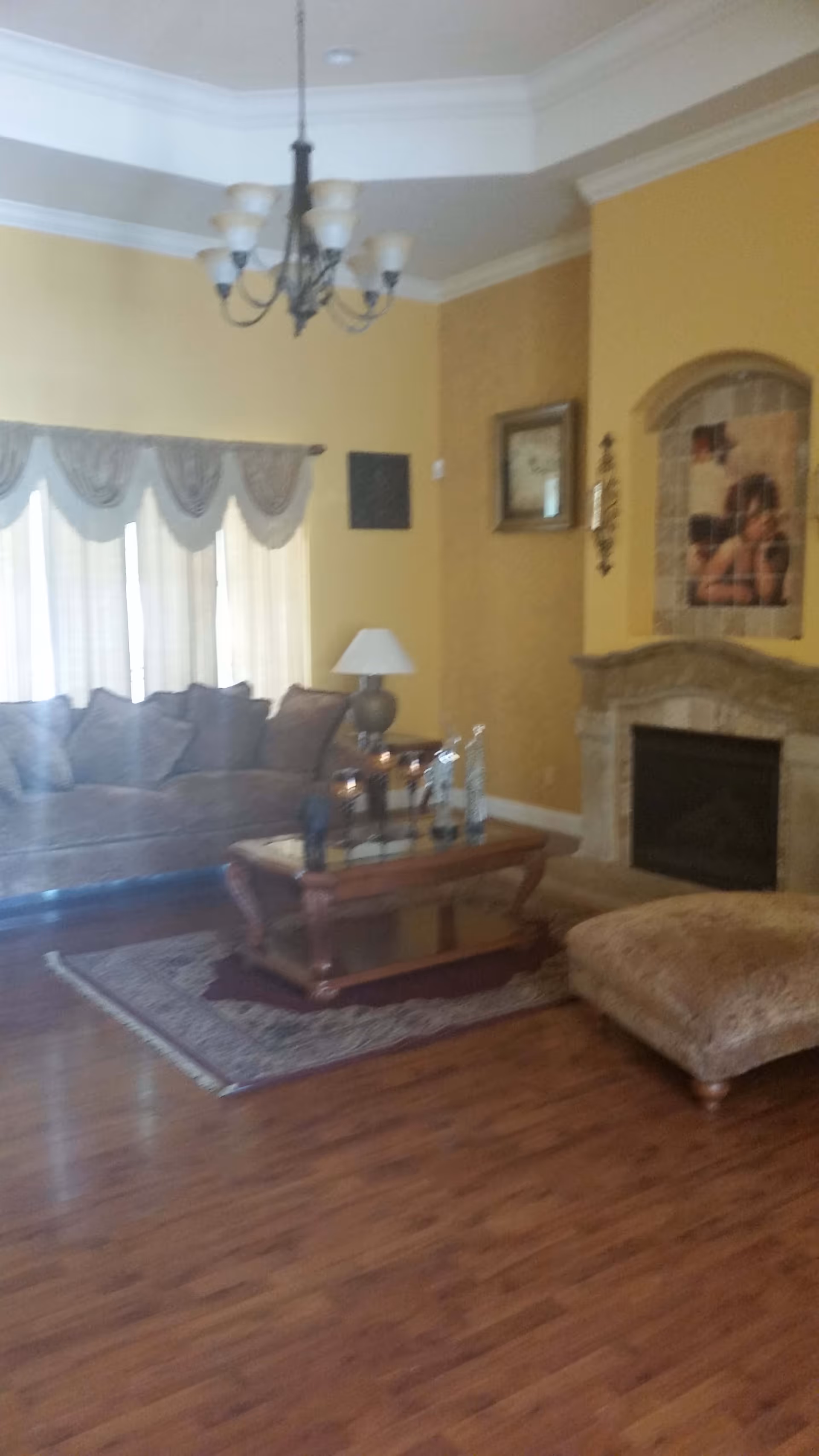 A cozy living room with a brown sofa, a wooden coffee table on a rug, a large cushioned ottoman, a table lamp, and a fireplace with a decorative tile inset. The walls are painted yellow and there is a chandelier hanging from the ceiling.