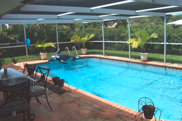A screened-in outdoor swimming pool area with clear blue water, surrounded by a brick border and patio. There are several potted plants along the pool edge and a black inflatable orca floating in the pool. Patio furniture including a table and chairs is visible on the left side.