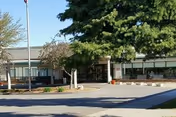 Exterior view of a single-story building with large windows and a covered entrance, surrounded by trees and landscaping under a clear sky.