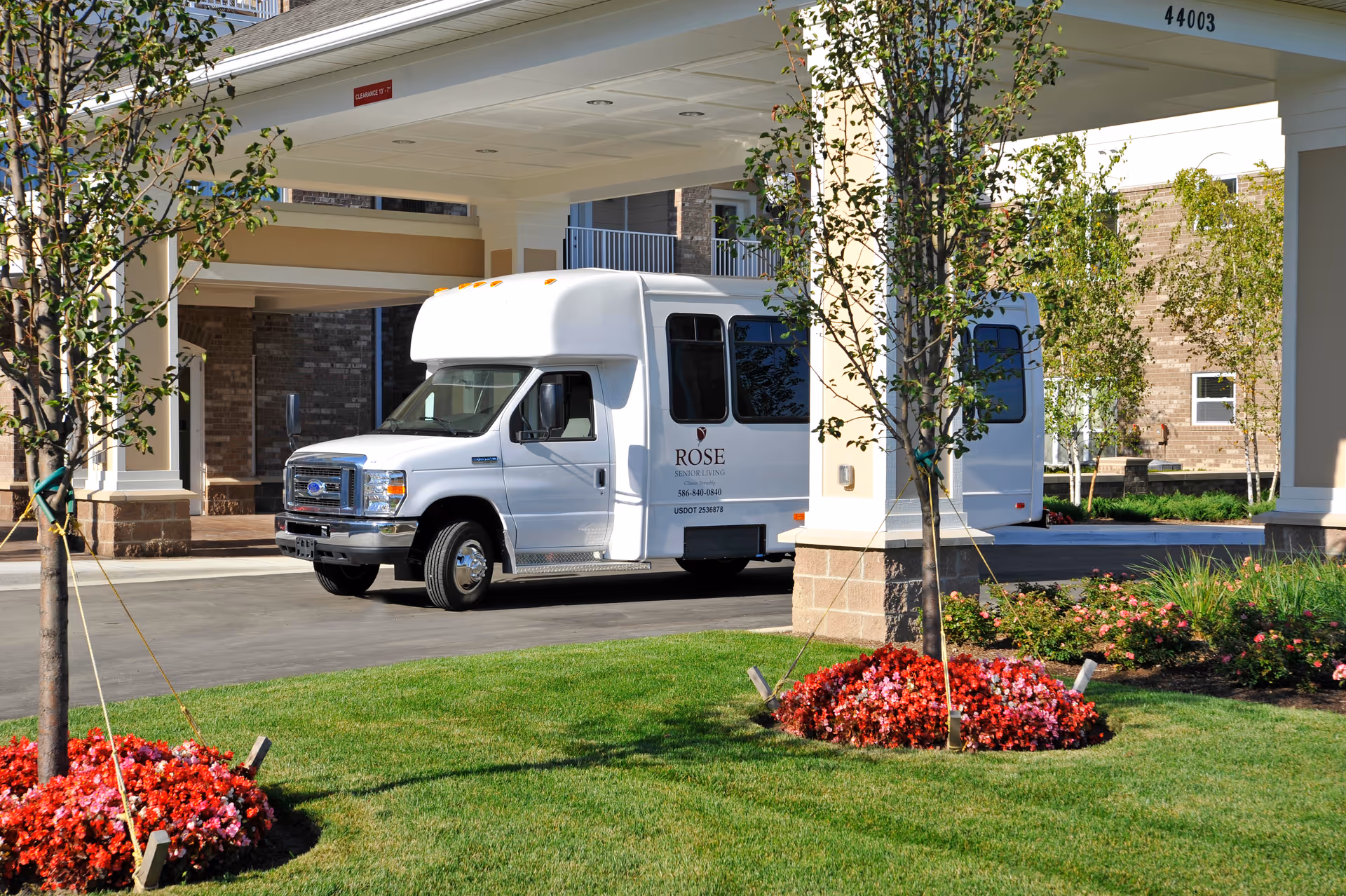 A white Rose Senior Living shuttle parked under the covered entrance of a senior living building with landscaped flower beds in front.
