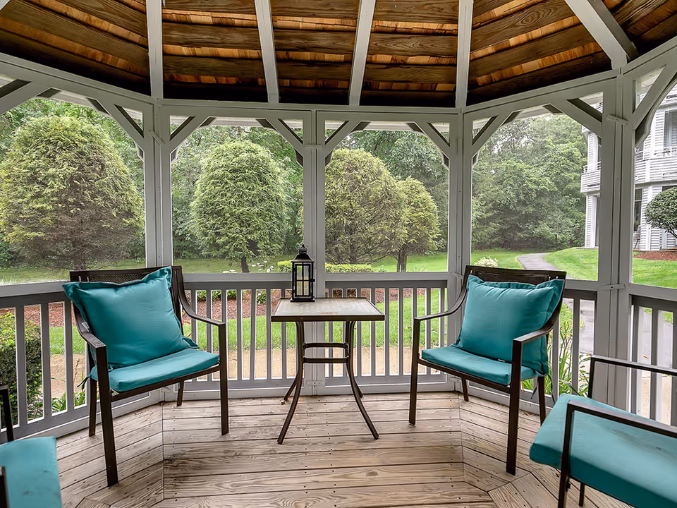 View from a wooden gazebo furnished with two teal-cushioned chairs, a small table with a lantern, and a leafy garden beyond.