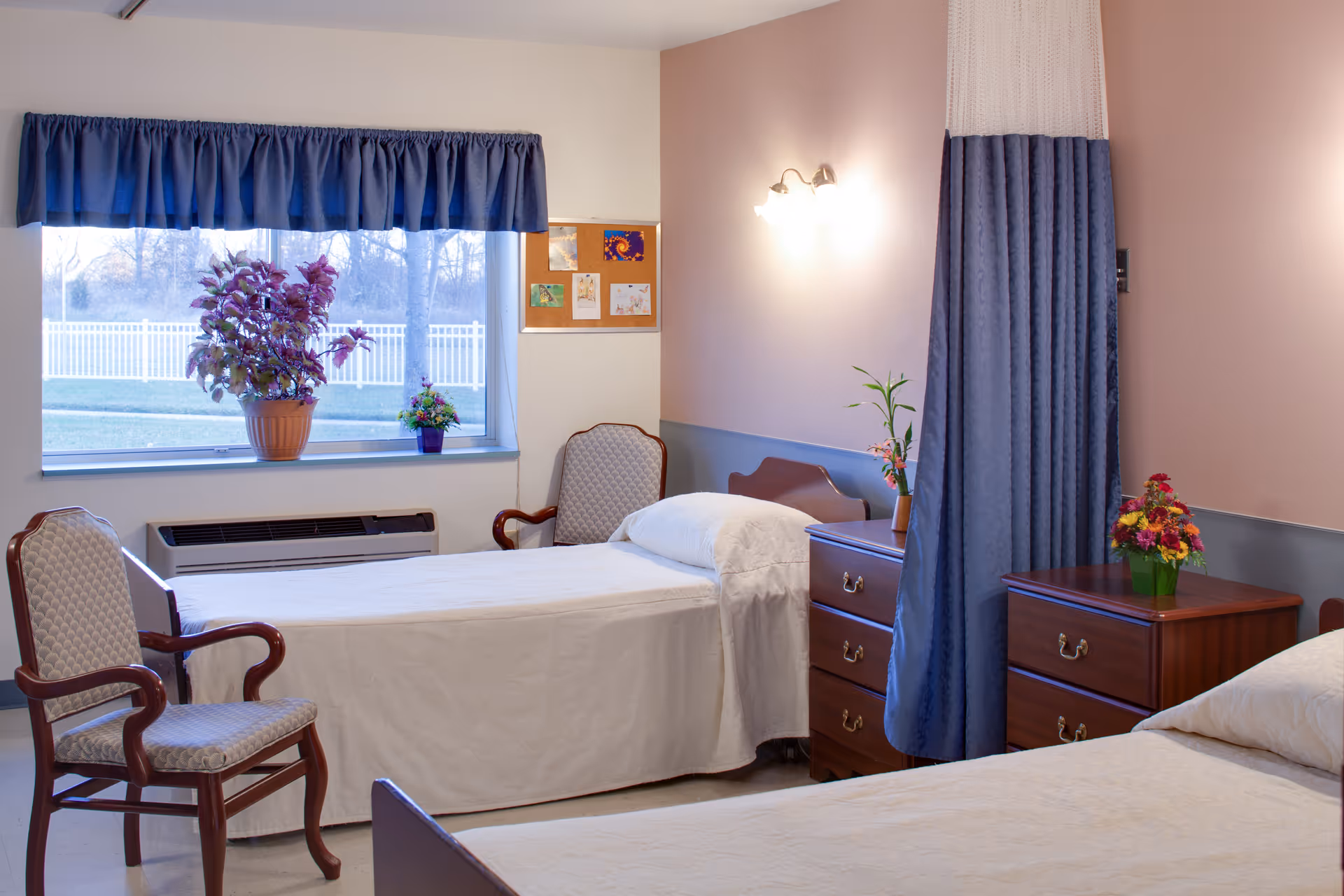 A well-lit bedroom in a senior living facility with two single beds covered in white linens. There are two wooden nightstands with drawers, each holding a small vase of flowers. A blue privacy curtain hangs between the beds. Two upholstered chairs with wooden arms are positioned near a window with blue valance curtains. Outside the window, a white fence and trees are visible.