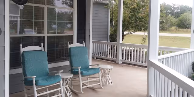 A covered porch area with two white rocking chairs featuring teal cushions and two small white side tables. The porch has white railings and overlooks a grassy yard with trees in the background.
