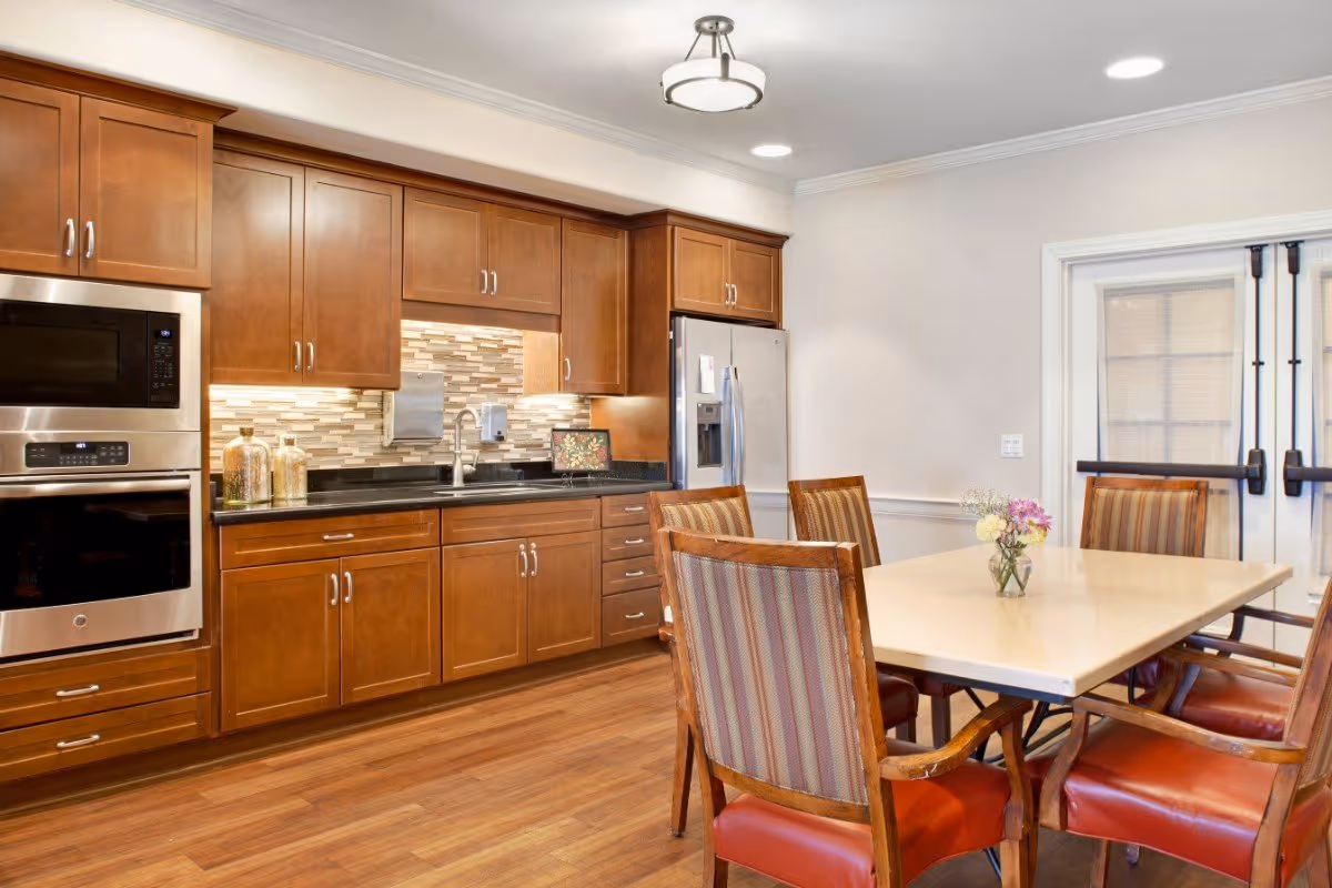 A kitchen and dining area with wooden cabinets, a stainless steel microwave and oven, a refrigerator, and a countertop with a sink. There is a dining table with six wooden chairs featuring striped upholstery and red leather seats. A small vase with flowers is placed on the table. The room has wooden flooring and white walls with a double glass door in the background.