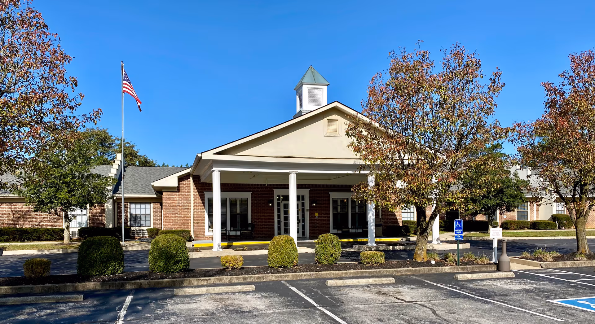 Front exterior view of a single-story brick building with a covered entrance supported by white columns. There are trimmed bushes and trees with autumn foliage in front of the building, an American flag on a flagpole to the left, and a clear blue sky overhead. The parking lot in front includes a handicapped parking space.